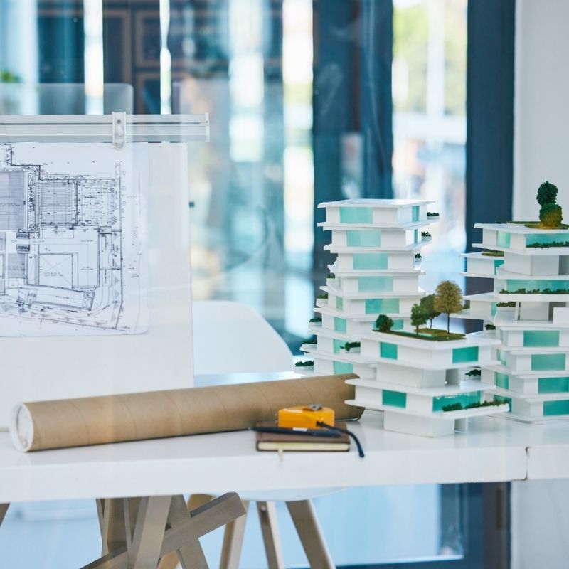 Architectural floor plans displayed on a drafting board next to a detailed building scale model on a white desk in a modern office