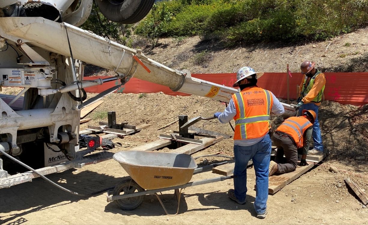 Close-up of workers guiding concrete from a truck chute into formwork for shoring caissons