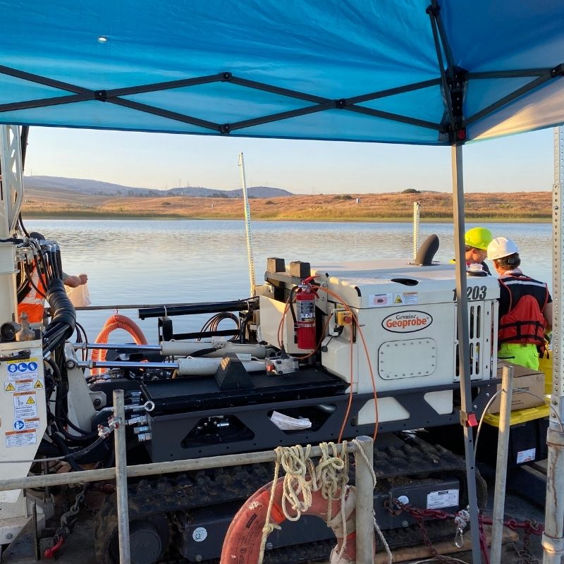Geotechnical drilling equipment mounted on a floating platform positioned over a calm reservoir