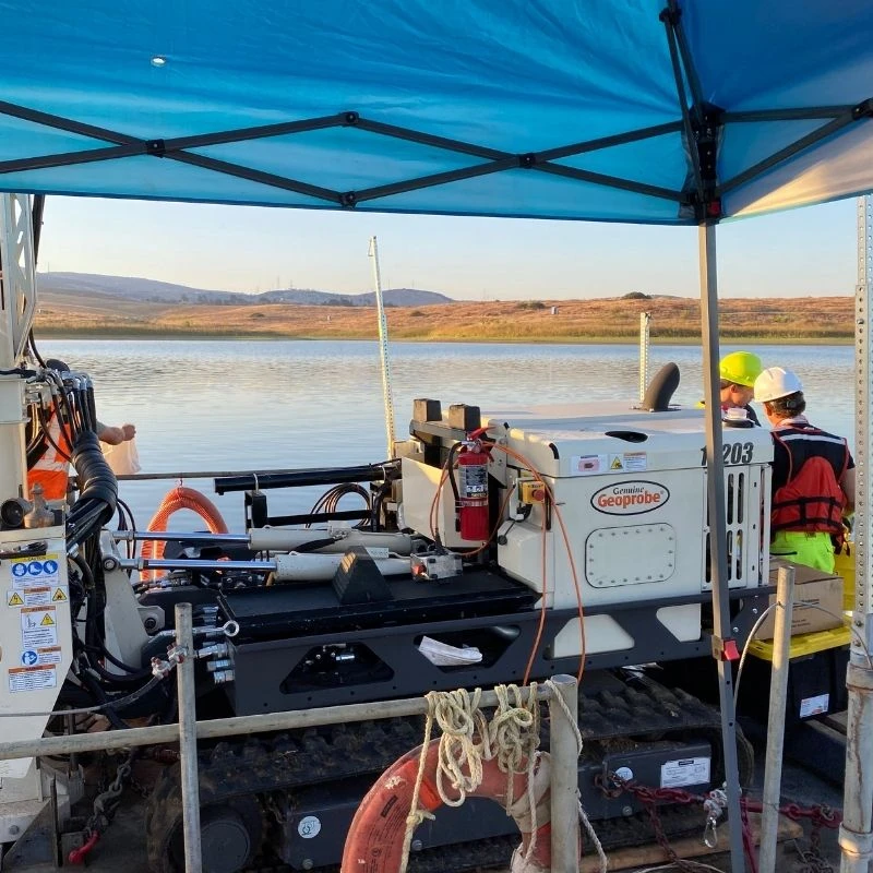 Geotechnical drilling equipment mounted on a floating platform positioned over a calm reservoir