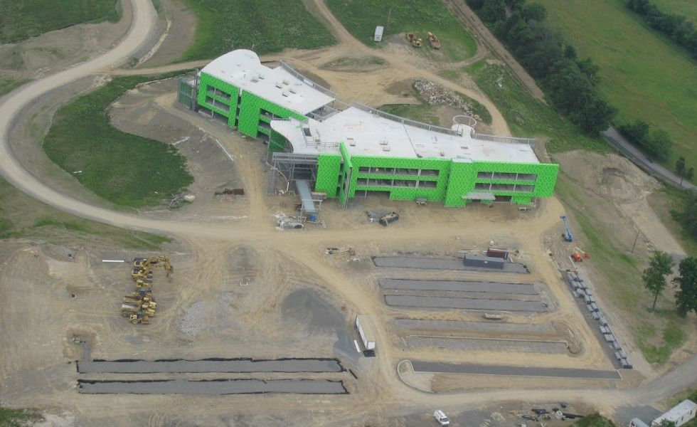Aerial view of the Mediacom Corporate Headquarters construction project in New York. The partially completed office building is surrounded by active site work, including grading, paved sections, heavy equipment, and temporary construction roads.