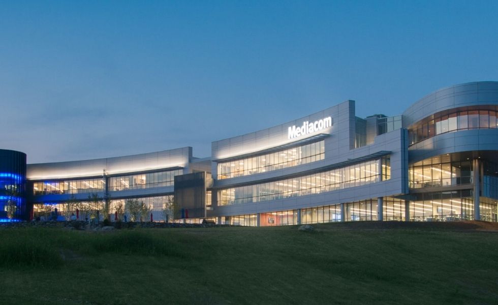 Evening exterior photograph of the Mediacom Corporate Headquarters in New York. The multi-story corporate office building is illuminated from within, highlighting its curved architecture, extensive glazing, and contemporary metal façade against a twilight sky.