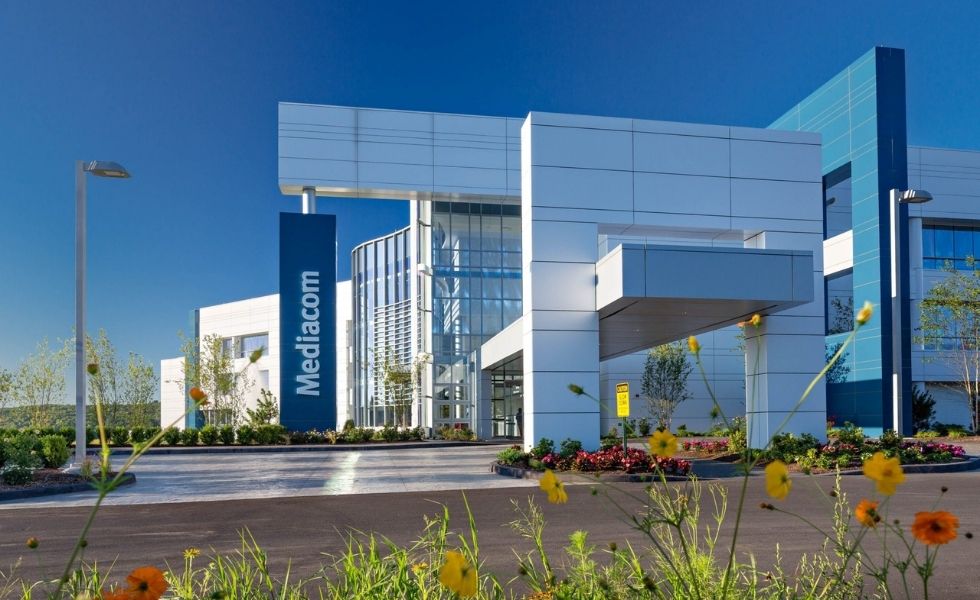 Front entrance of the Mediacom Corporate Headquarters in New York featuring a contemporary architectural design with metal panel cladding, expansive glass walls, a covered drop-off canopy, landscaped flower beds, and prominent Mediacom signage at the building entrance.