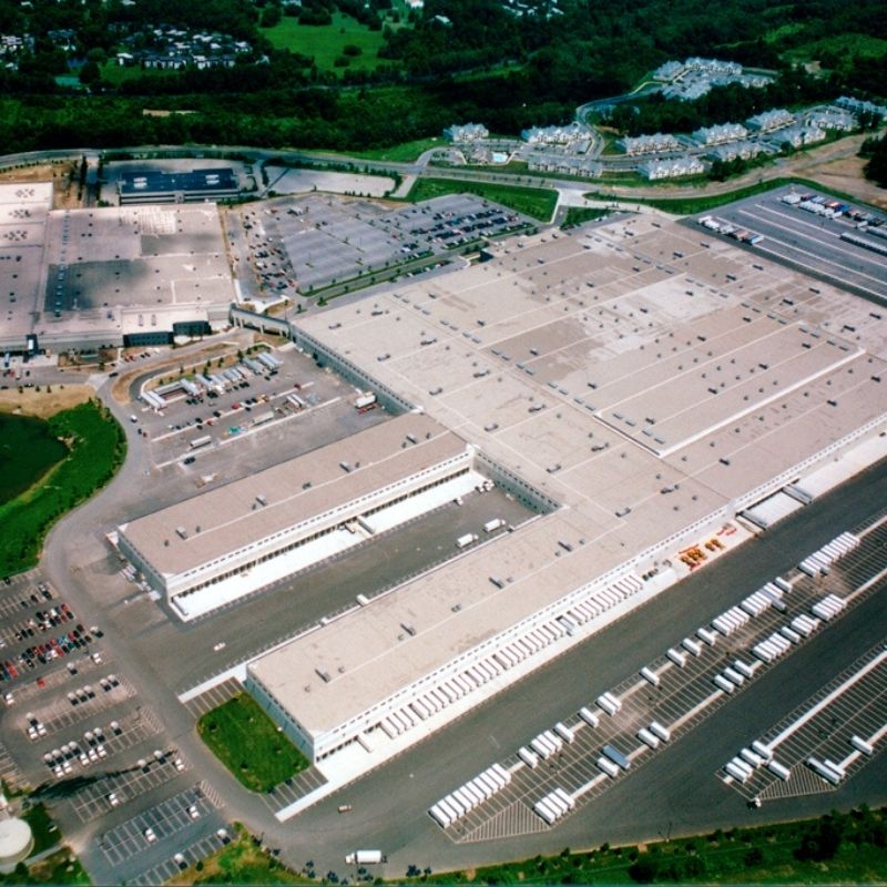 Aerial view of a large distribution and logistics facility with expansive warehouse buildings, multiple loading docks, truck parking areas, employee parking lots, access roads, and surrounding green spaces.