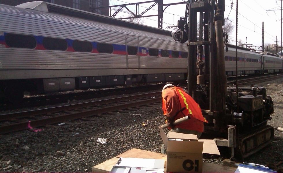 41st Street Bridge project: geotechnical drilling operations underway adjacent to active railway tracks, with a passenger train passing through the project area.