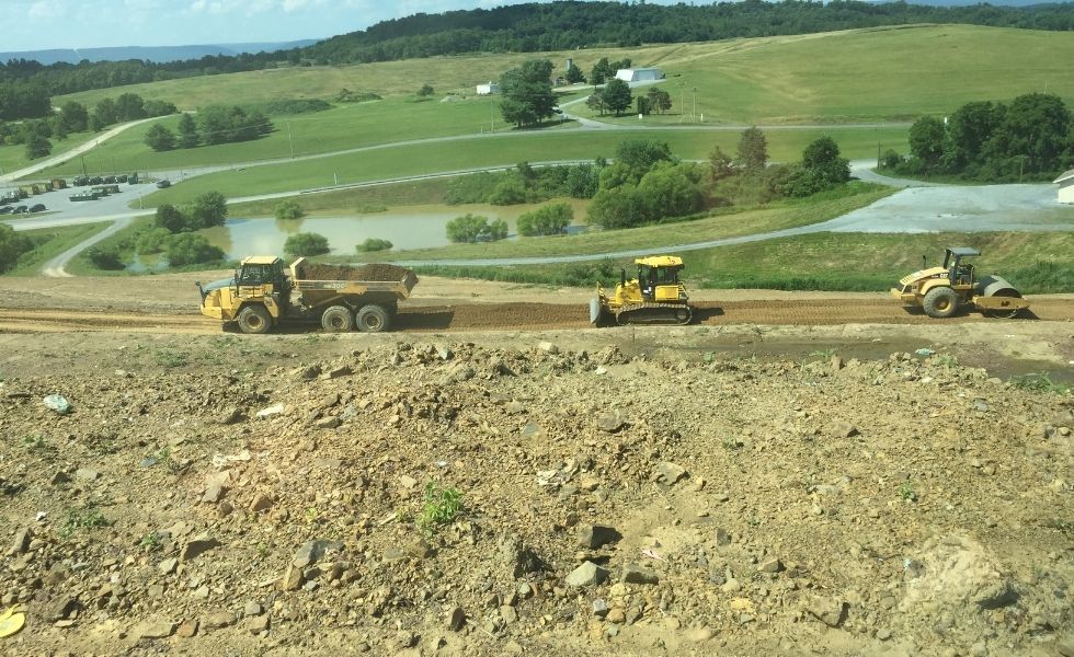 Dump truck, bulldozer, and compactor operating on a graded construction site with rolling terrain and stormwater features in the background at the PhilaPort Auto Storage Facility.