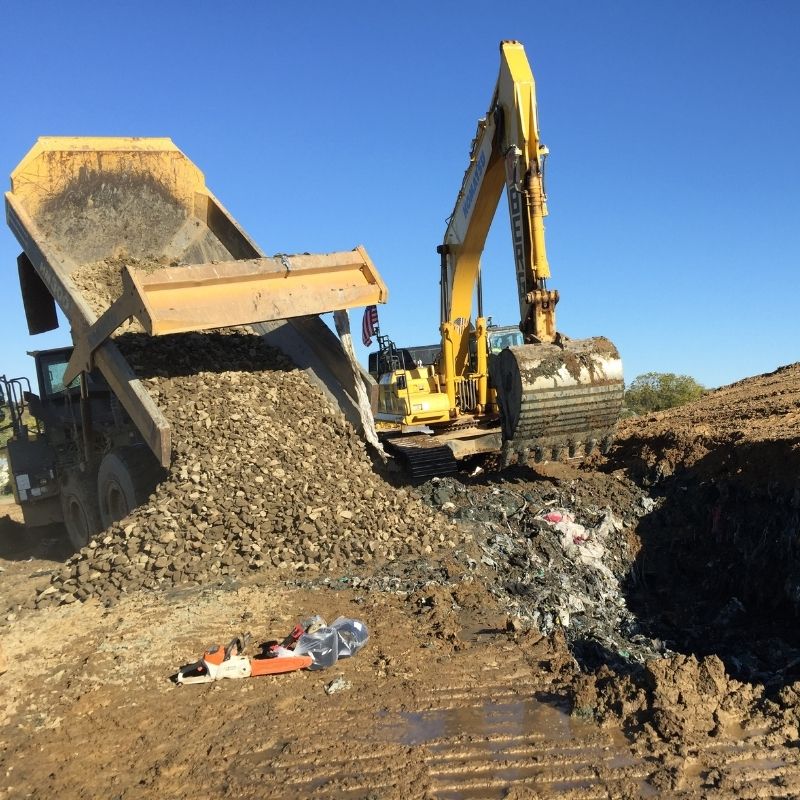 Heavy equipment operating at the Lebanon Landfill project, with a dump truck unloading aggregate material beside a large excavator working in an excavation trench.
