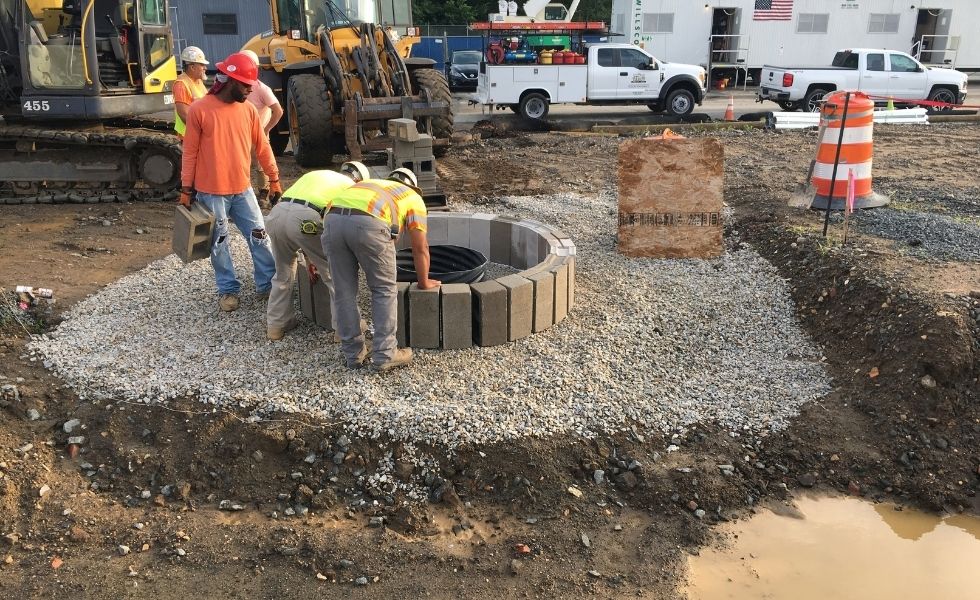 Construction crew installing a circular precast stormwater drainage structure on a gravel base at the Philaport Auto Facility project site, with heavy equipment