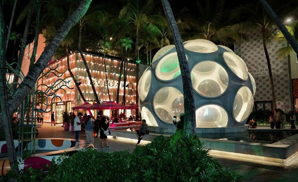 An evening view of the Miami Design District featuring an illuminated spherical pavilion surrounded by palm trees, luxury retail storefronts