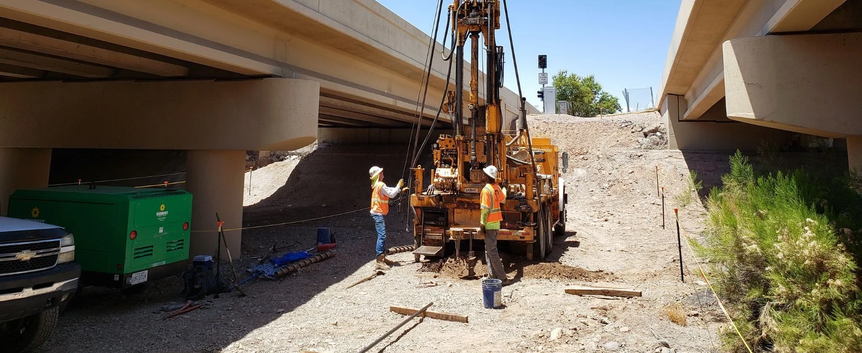 Construction workers operate a drilling rig beneath a highway overpass
