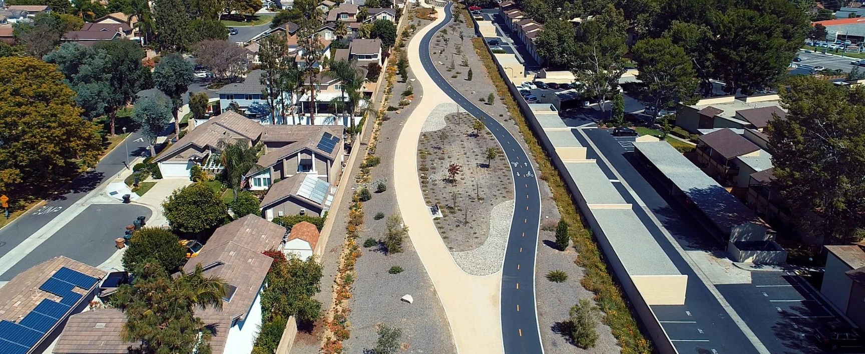 Aerial view of a winding paved bike path and walking trail running through a landscaped greenway between residential houses