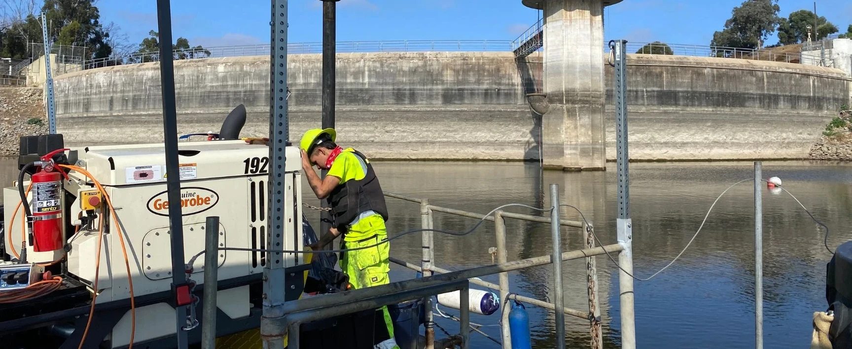 A field technician wearing high-visibility safety gear works on monitoring equipment next to a water basin at a concrete reservoir facility