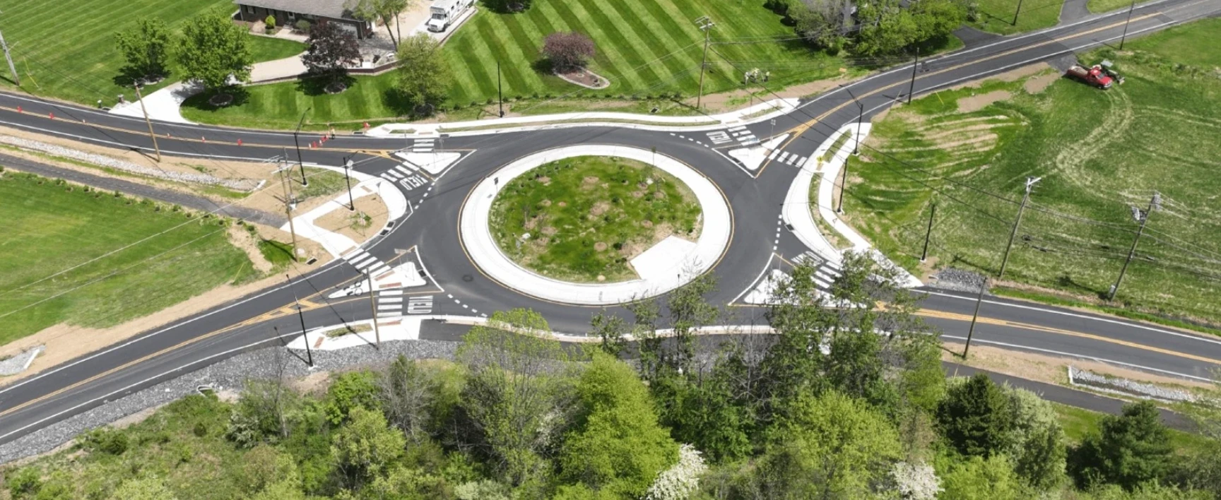 Aerial view of the Old Bethlehem Pike/Portzer Road Roundabout, showing the completed multi-lane roadway configuration, landscaped central island, pedestrian crosswalks, sidewalks, and surrounding residential and agricultural areas.