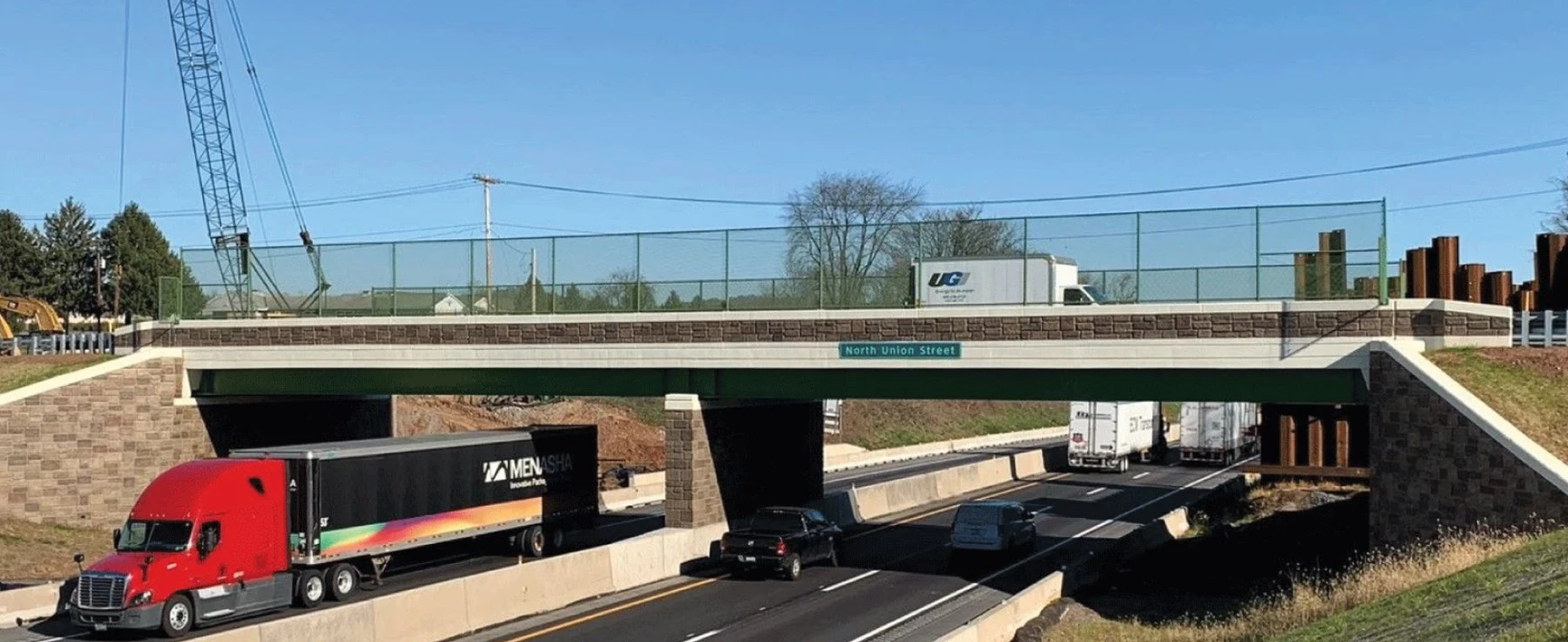 Completed highway overpass carrying local traffic above a multi-lane roadway as part of the PTC Bridge EB311 project, with bridge abutments, safety barriers, and vehicles traveling beneath the structure.