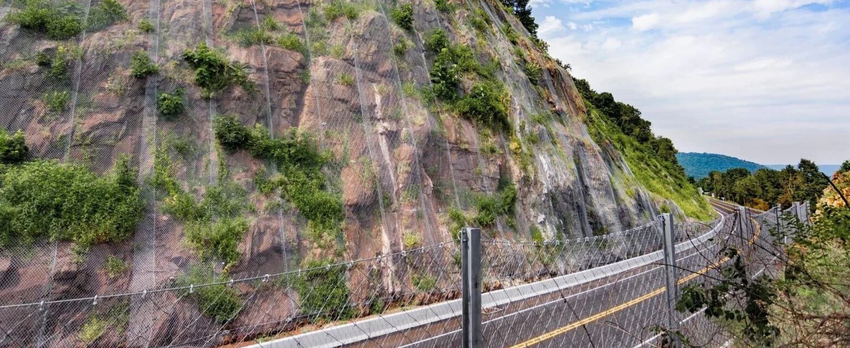Rockfall protection mesh covering a steep rock face adjacent to a roadway as part of the US 11/15 Rock Slope Safety Improvement project, with safety barriers and vegetated rock slopes visible along the transportation corridor.