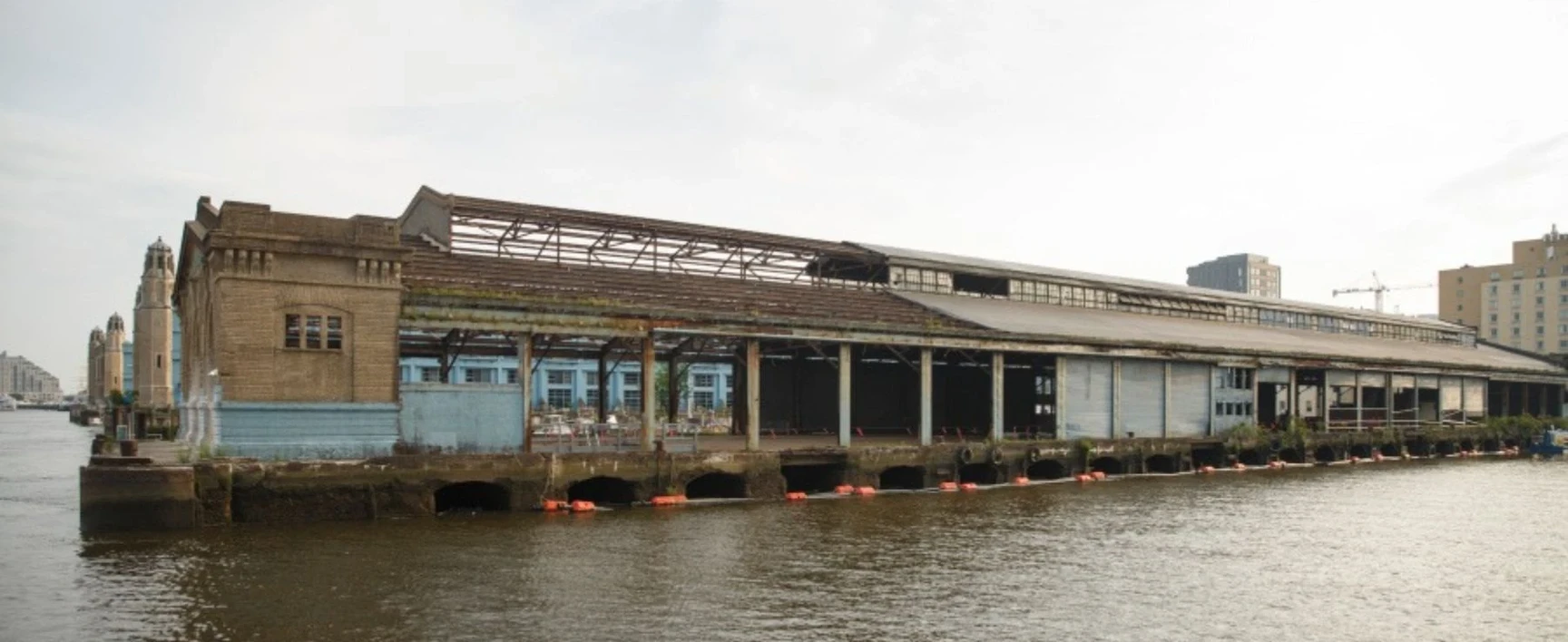 Existing waterfront warehouse structure at PhilaPort Pier 82S, showing the pier-supported building, loading bays, deteriorated roof framing, and marine infrastructure along the river prior to rehabilitation.