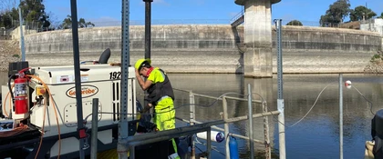 A field technician wearing high-visibility safety gear works on monitoring equipment next to a water basin at a concrete reservoir facility