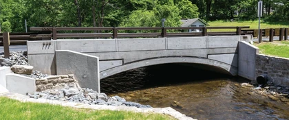 Completed McMichael’s Creek Bridge project in Pennsylvania, featuring a concrete arch bridge spanning a shallow stream, with roadway guardrails, stone riprap, and restored stream banks designed to improve transportation infrastructure and waterway resilience.